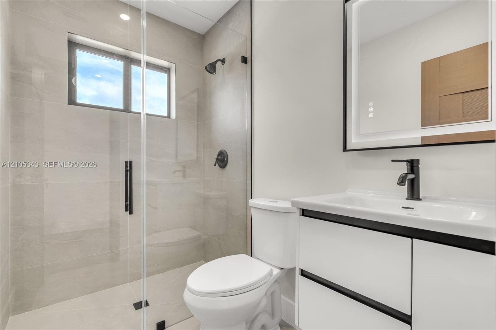 Modern bathroom featuring a frameless glass-enclosed shower, matte black plumbing fixtures, a white vanity with integrated basin, and a backlit wall-mounted mirror