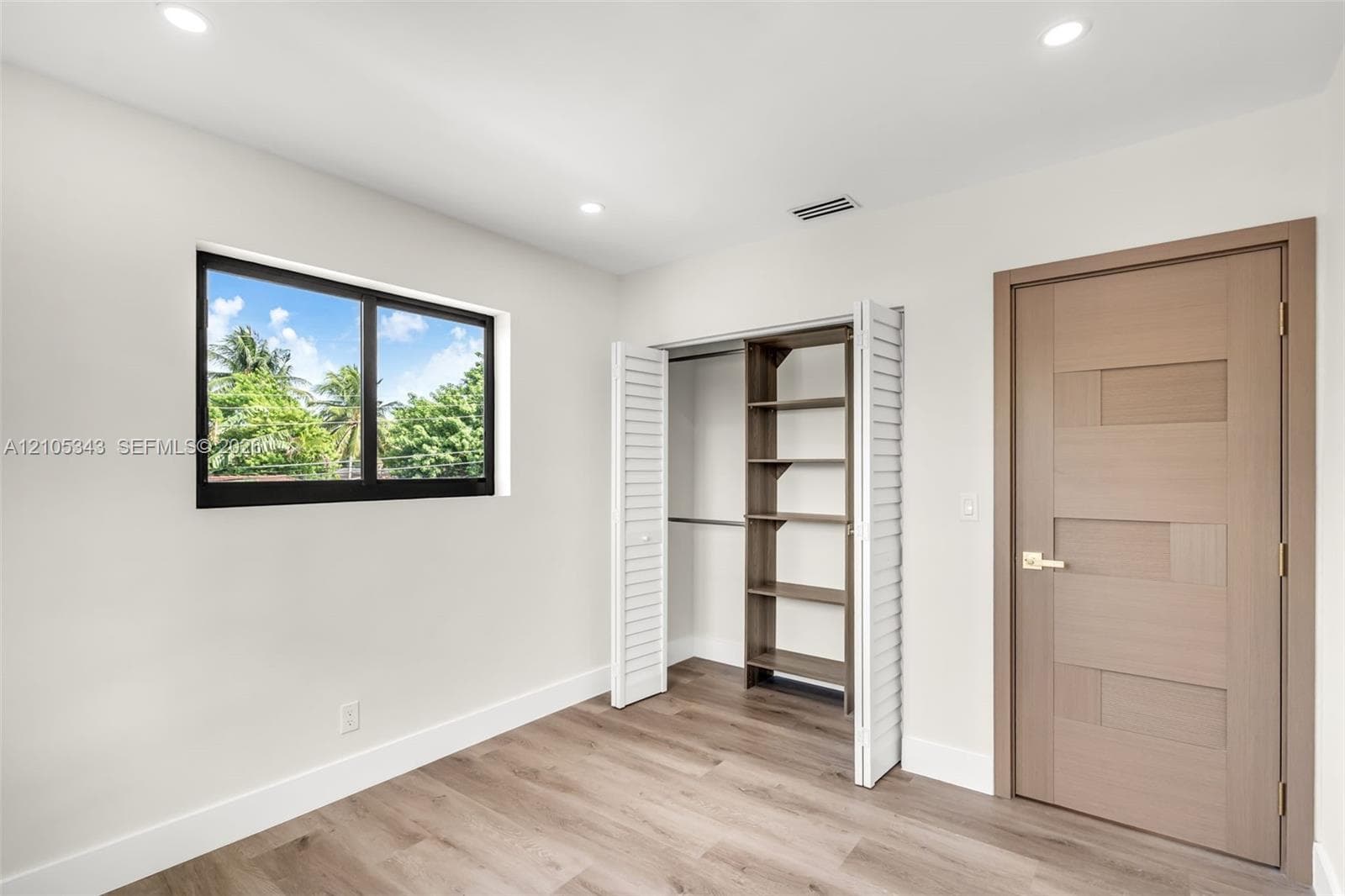Interior space featuring wood-finish flooring, recessed lighting, a single-pane window with black framing, a closet with built-in shelving, and a wood-textured door with gold-toned hardware