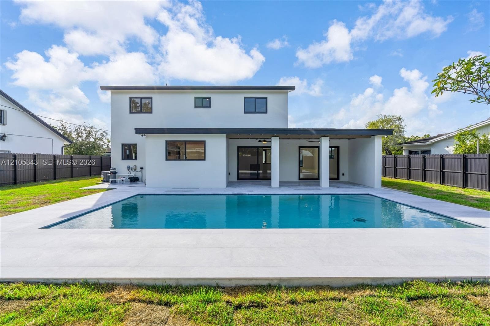 Rectangular in-ground pool with concrete surround, covered patio, smooth stucco exterior, and black perimeter fencing