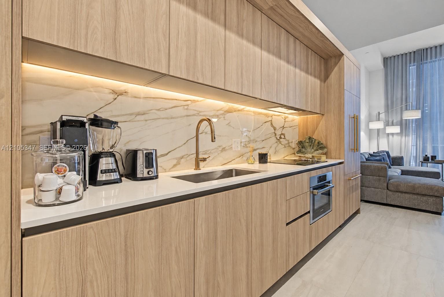 Integrated kitchen featuring light wood-finish cabinetry, under-cabinet lighting, a built-in oven, and a gold-tone faucet