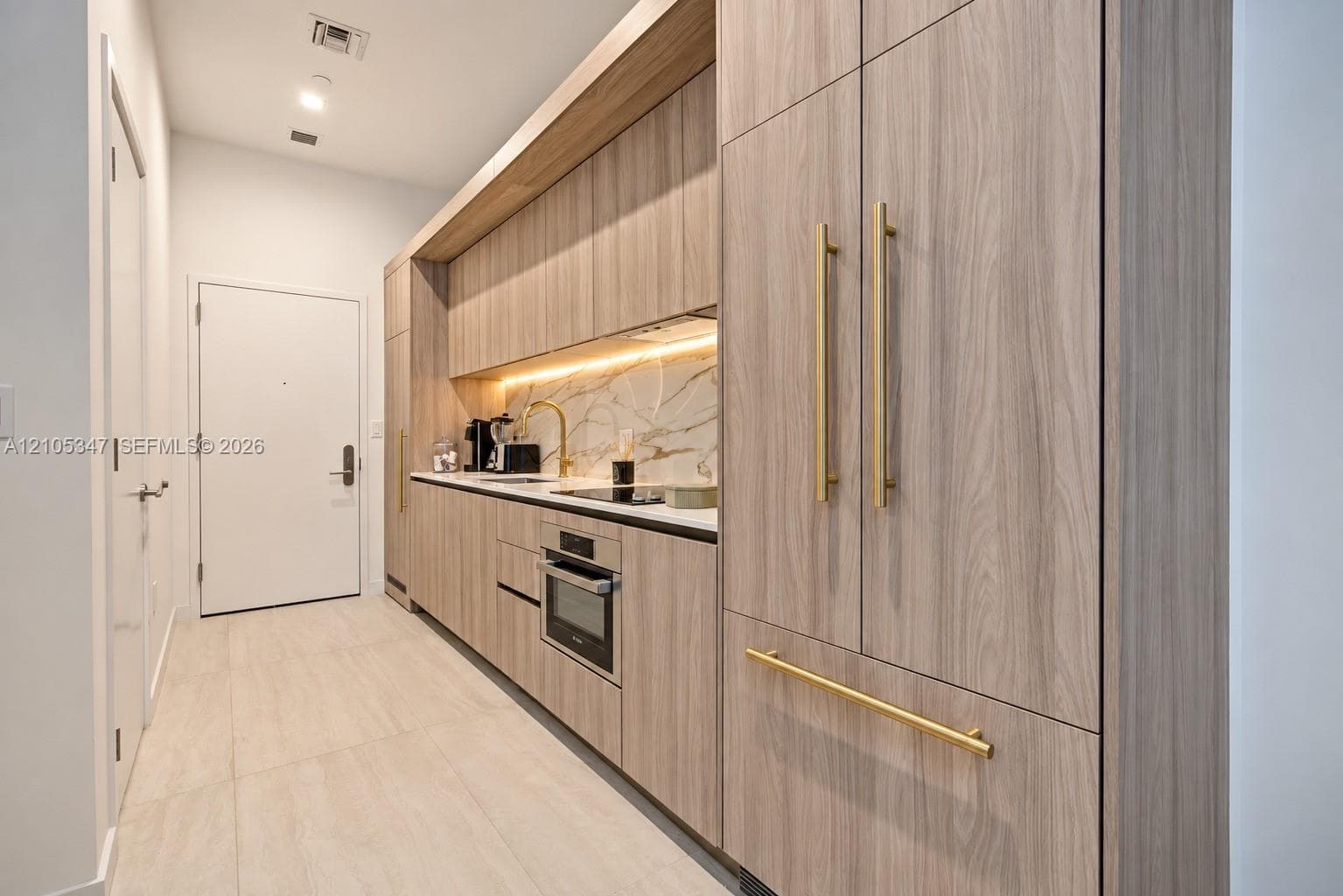 Full-height light wood-finish cabinetry with gold-tone hardware, integrated oven, under-cabinet lighting, and a white countertop with a matching backsplash