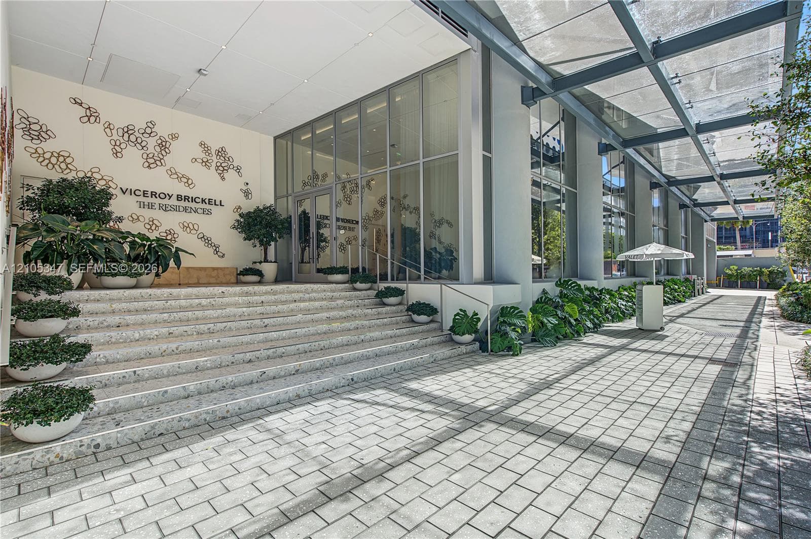 Building exterior featuring a glass curtain wall, expansive paver walkway, overhead steel-frame canopy, and integrated wide-tread stairs with decorative stonework