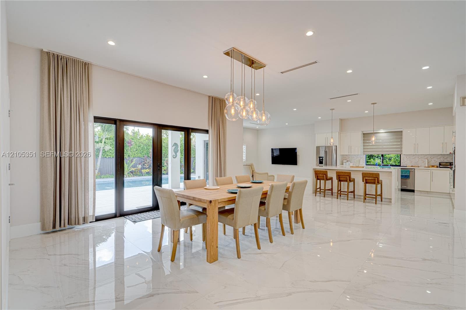 Open-concept interior featuring large-format polished stone flooring, recessed lighting, a modern multi-pendant chandelier, white slab-front cabinetry, and glass sliding doors