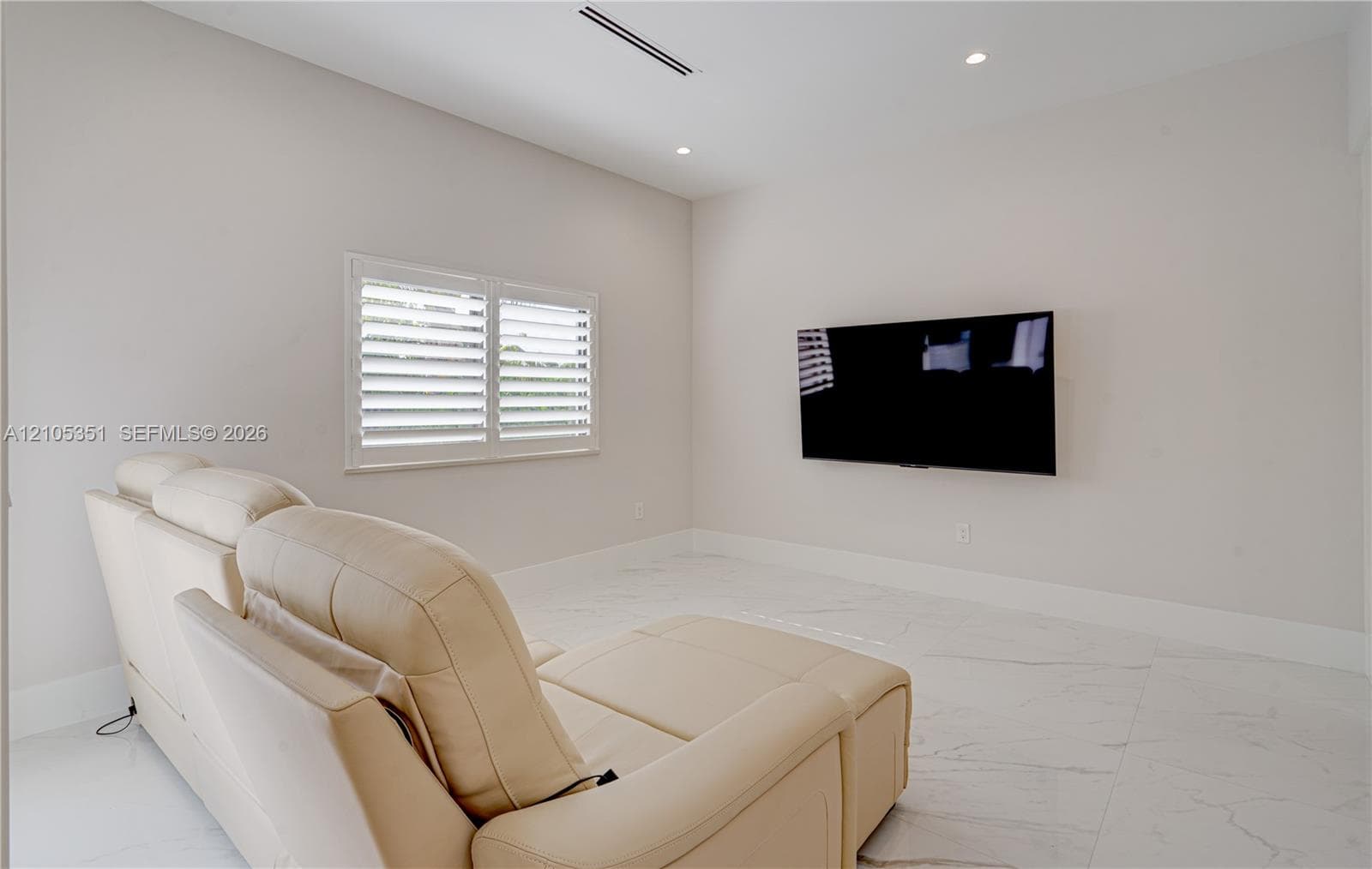 Neutral-toned interior featuring large-format tile flooring, recessed ceiling lighting, a wall-mounted linear HVAC vent, and a window fitted with white plantation shutters
