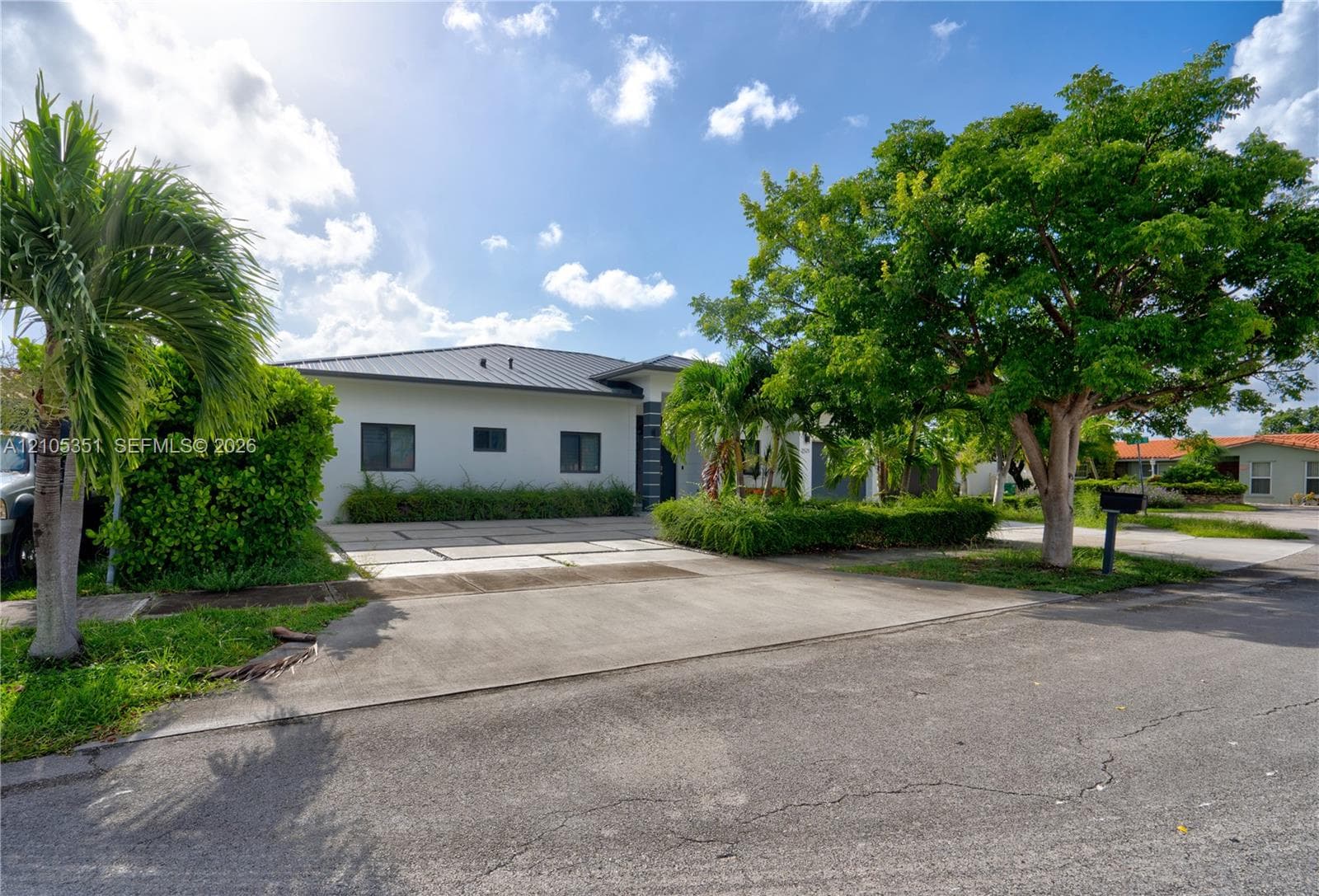 Single-story exterior featuring a metal roof, smooth stucco facade, expansive paved driveway, and mature landscaping