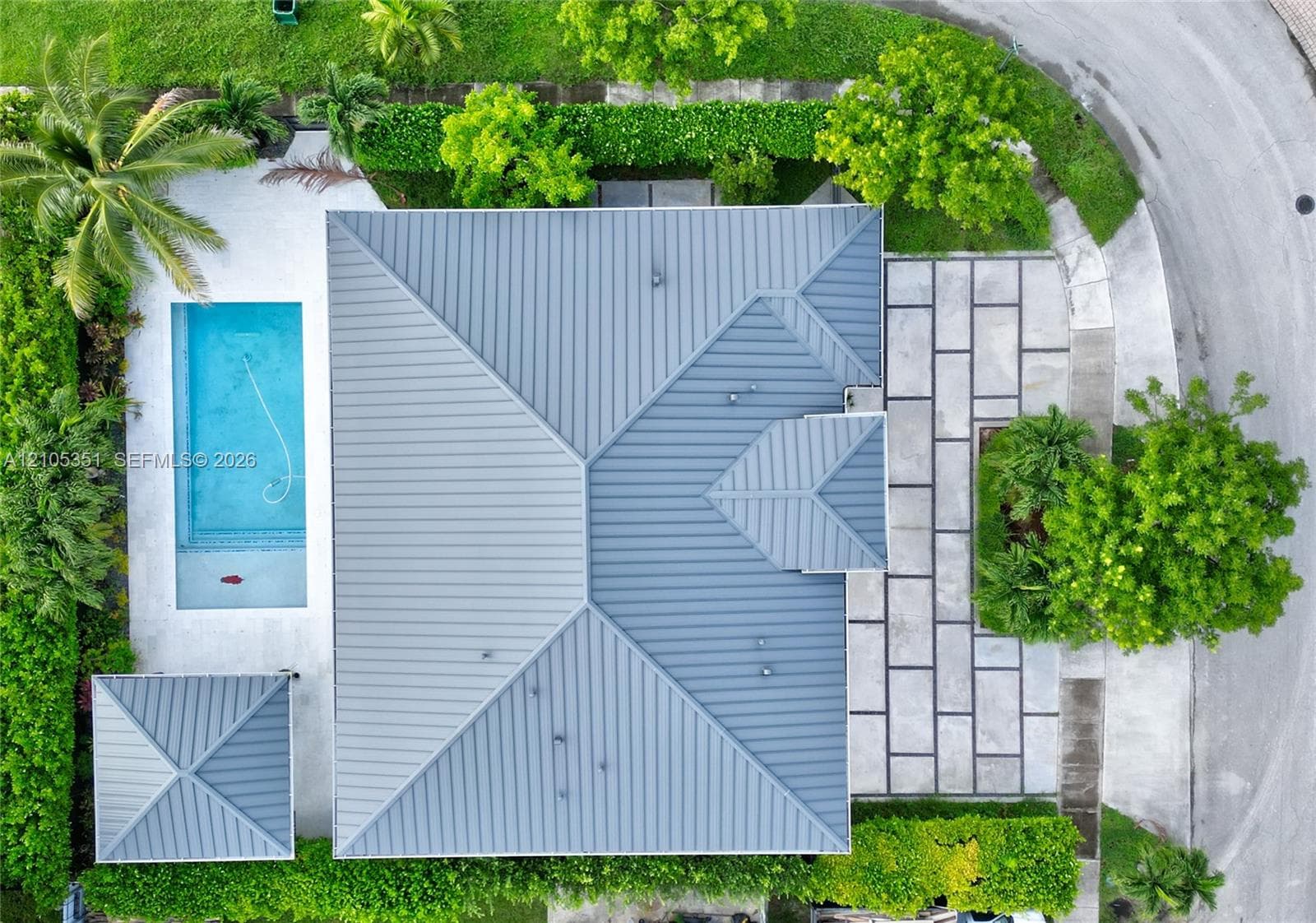 Aerial perspective featuring a metal roof, rectangular swimming pool, separate cabana structure, and expansive stone paver hardscaping bordered by mature hedges