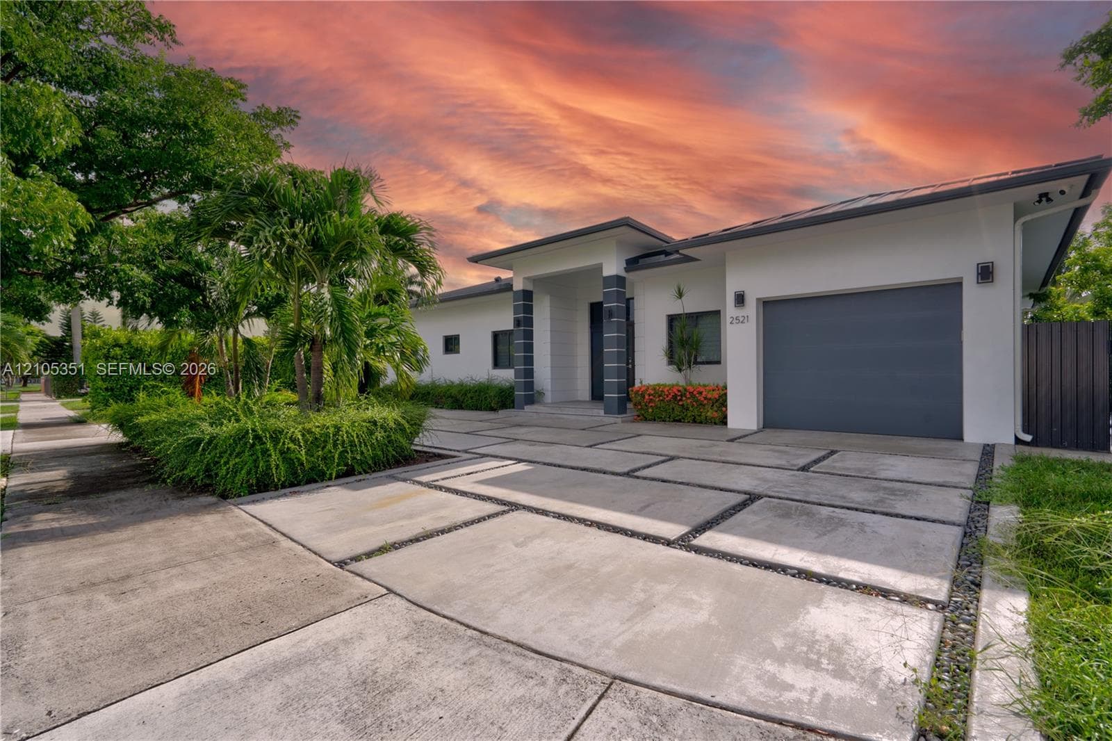 Modern single-story exterior featuring a geometric concrete paver driveway, covered entryway with column detailing, attached garage, and mature professional landscaping