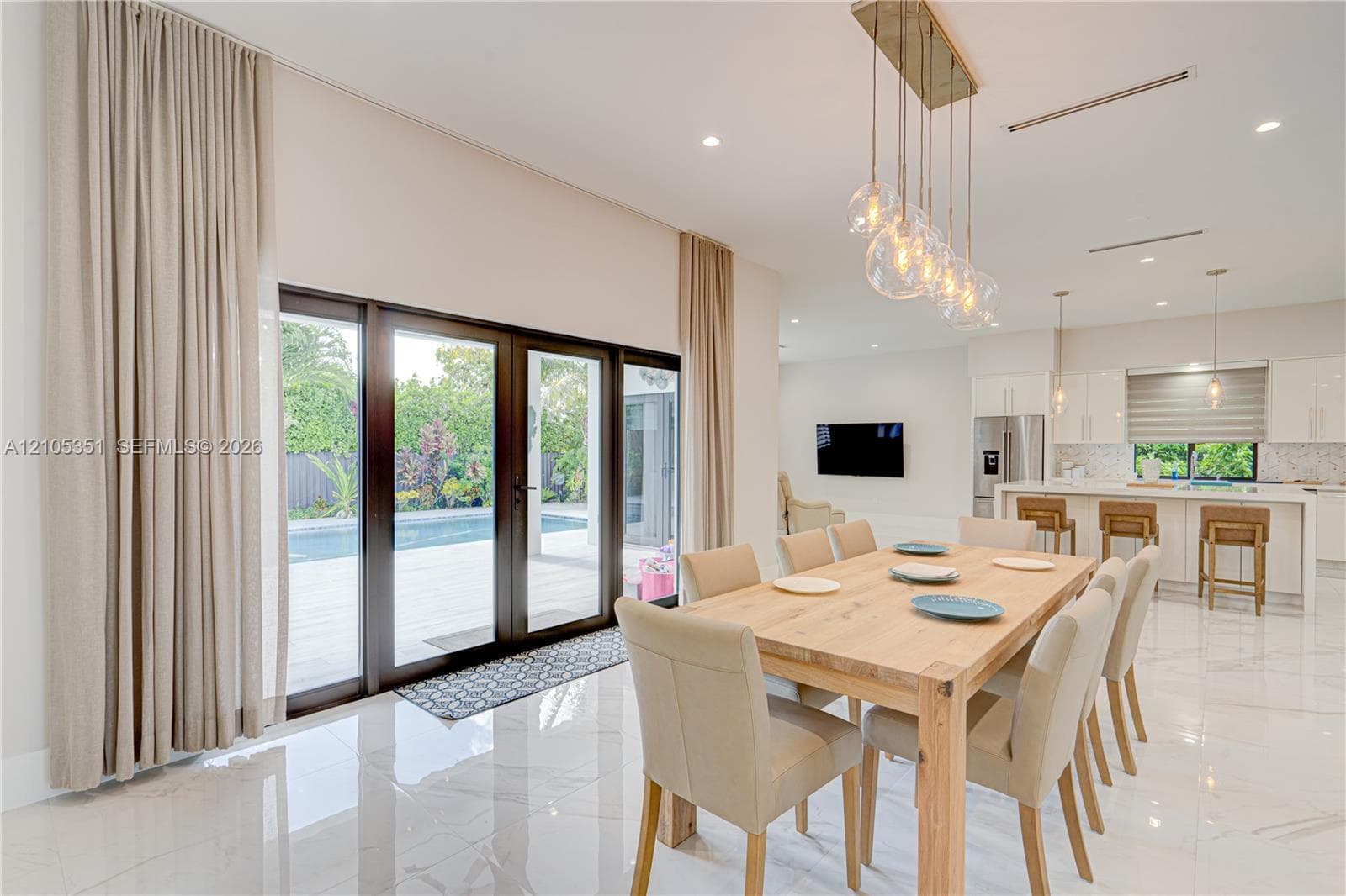 Open-concept interior featuring large-format polished tile flooring, floor-to-ceiling glass panel doors, a multi-light contemporary glass chandelier, and white cabinetry with a quartz waterfall-edge island