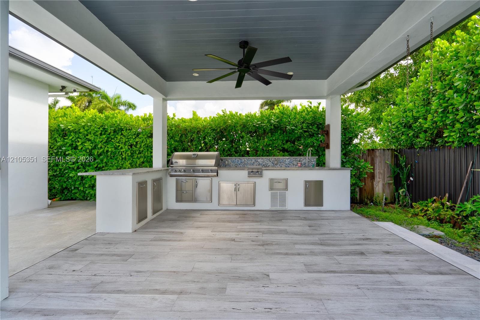 Covered outdoor kitchen featuring wood-finish tile flooring, built-in stainless steel grill, stone-style countertop, mosaic tile backsplash, and overhead ceiling fan