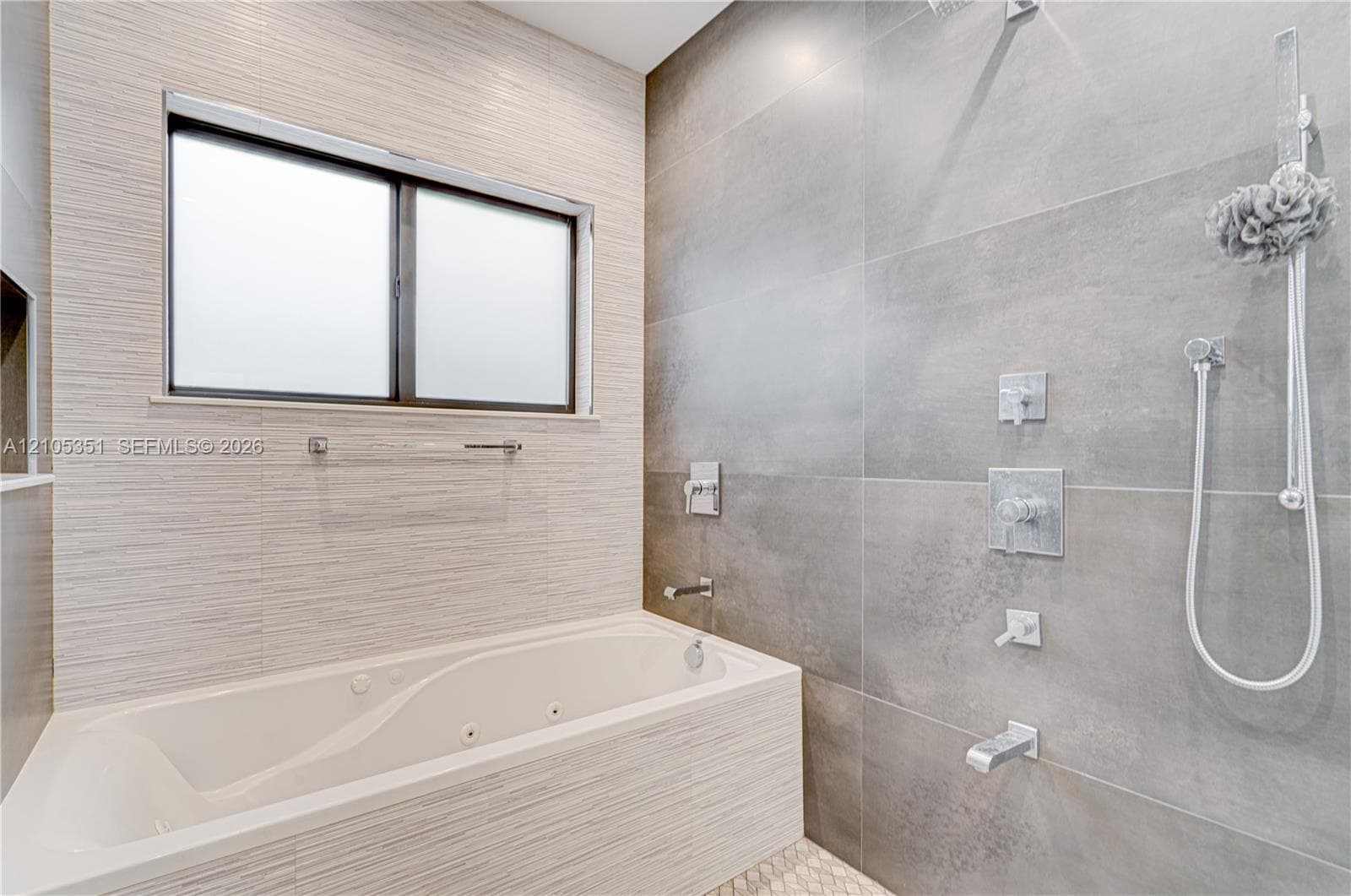 Jetted bathtub featuring textured tile surround, large-format grey wall tile, chrome shower fixtures with handheld sprayer, and rectangular window