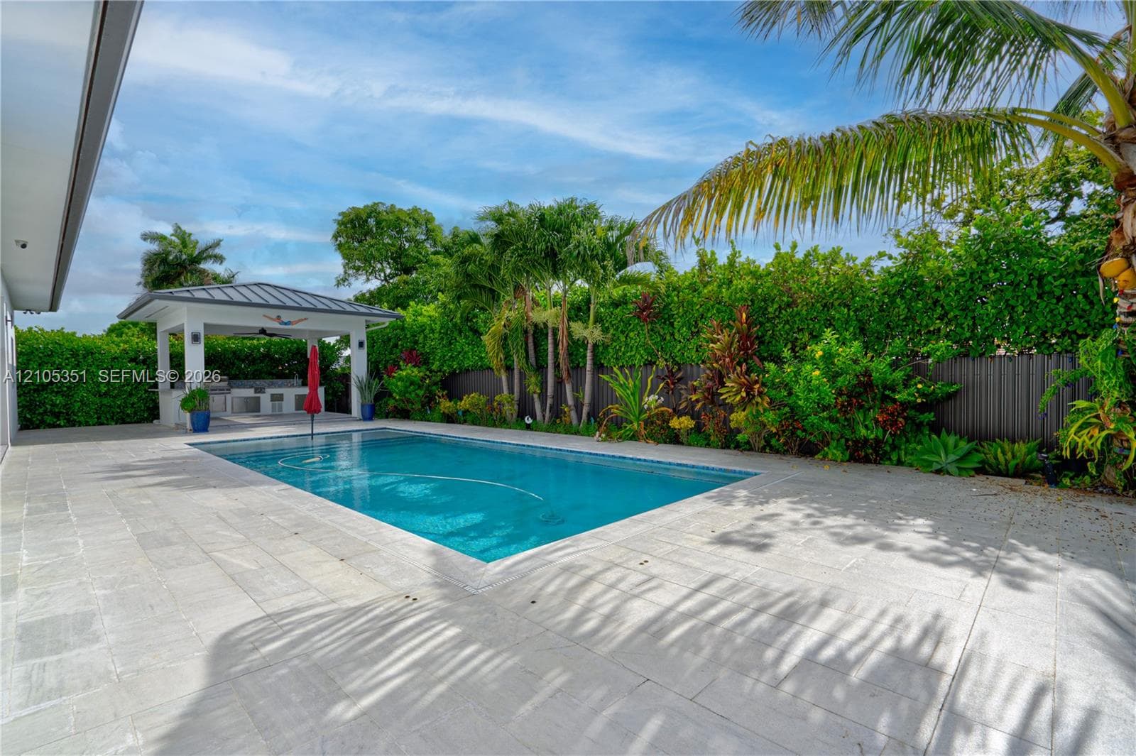 In-ground swimming pool with stone-tiled decking and a covered outdoor kitchen featuring built-in cabinetry and professional-grade appliances, enclosed by mature perimeter landscaping