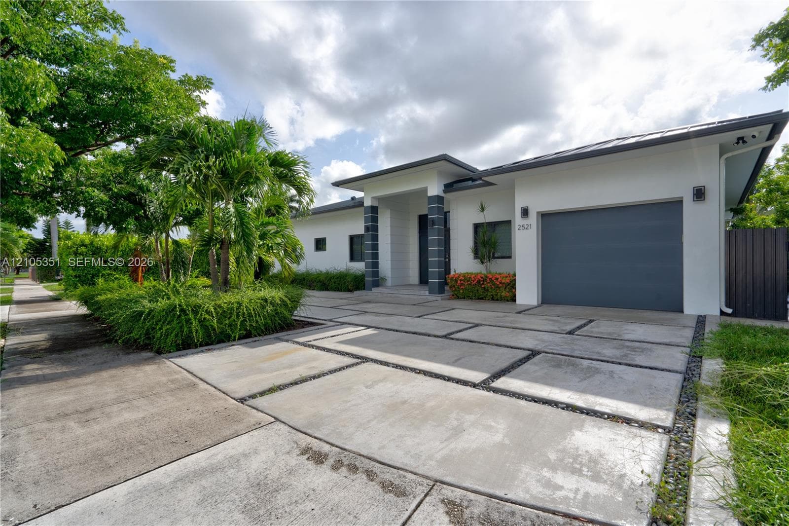 Contemporary single-story exterior featuring white stucco siding, a charcoal-toned garage door, geometric concrete paver driveway with decorative stone accents, and columns with tile detailing