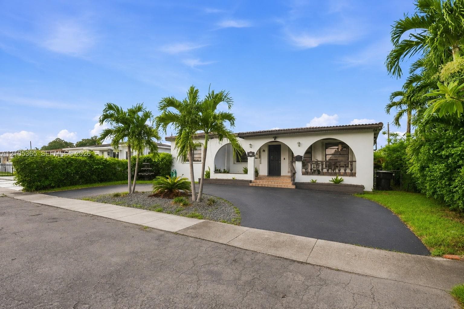 White stucco exterior with arched entryways, dark tile roof, paved driveway, and mature palm trees