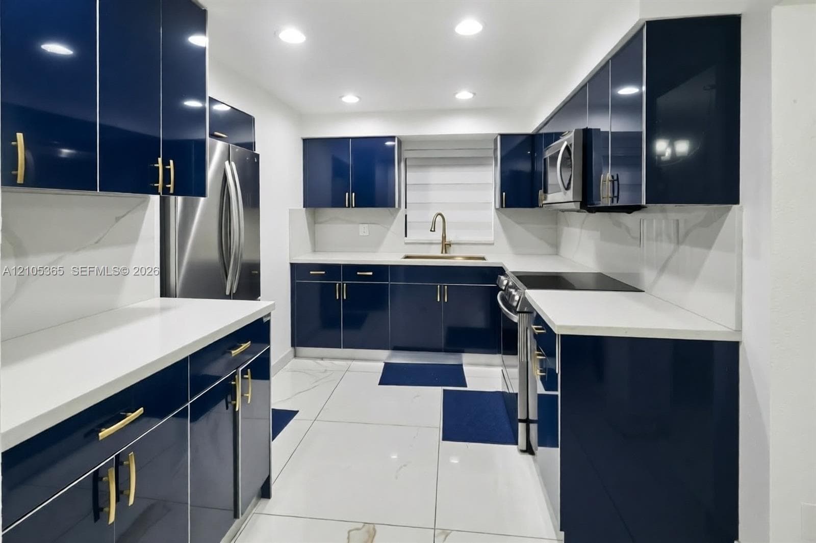 Glossy navy blue cabinetry with gold-tone hardware, white countertops, stainless steel appliances, and white tile flooring