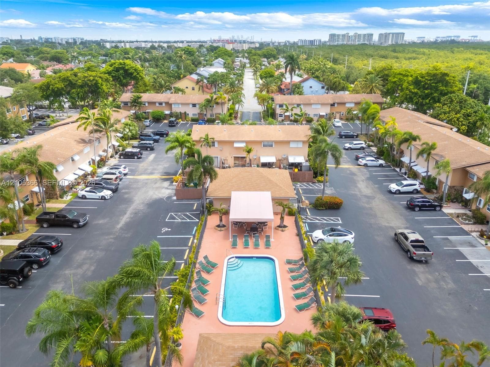 Central swimming pool with lounge area, surrounded by multiple residential buildings with ample parking, and mature palm trees