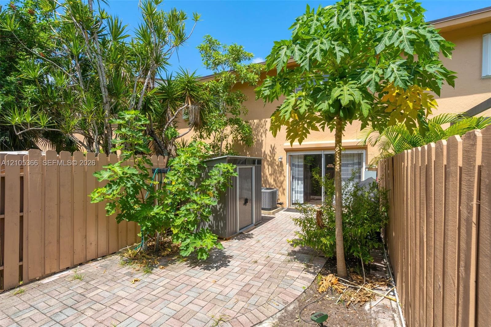 Paved outdoor area featuring a sliding glass door, privacy fencing, and tropical landscaping