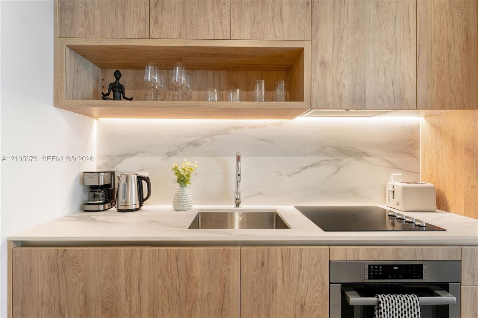 Light wood-finish cabinetry with an open shelf, undermount stainless steel sink, integrated cooktop, stainless steel built-in oven, and white marble-patterned backsplash