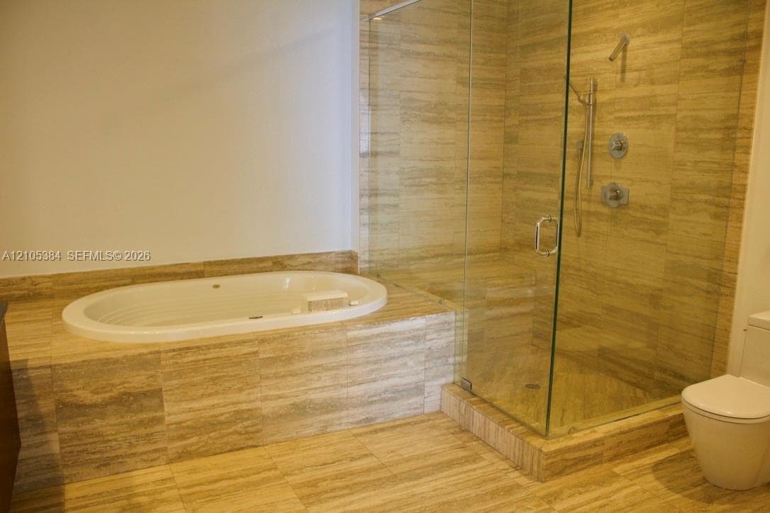 Recessed soaking tub, glass-enclosed shower with a handheld shower fixture, and a toilet, all set within a natural stone-tiled floor and wall surround