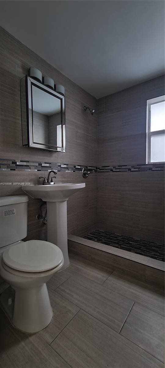 Pedestal sink with chrome faucet, built-in medicine cabinet with integrated lighting, tiled shower with textured floor, full wall tiling with decorative accent band, and a window for natural light