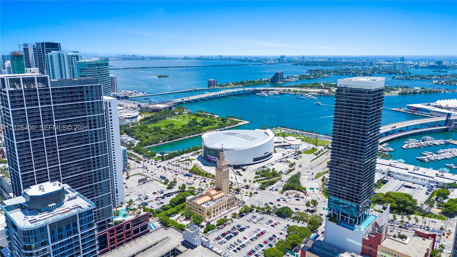 Expansive cityscape featuring high-rise buildings, a prominent arena structure, a large body of water, and a marina with multiple boats