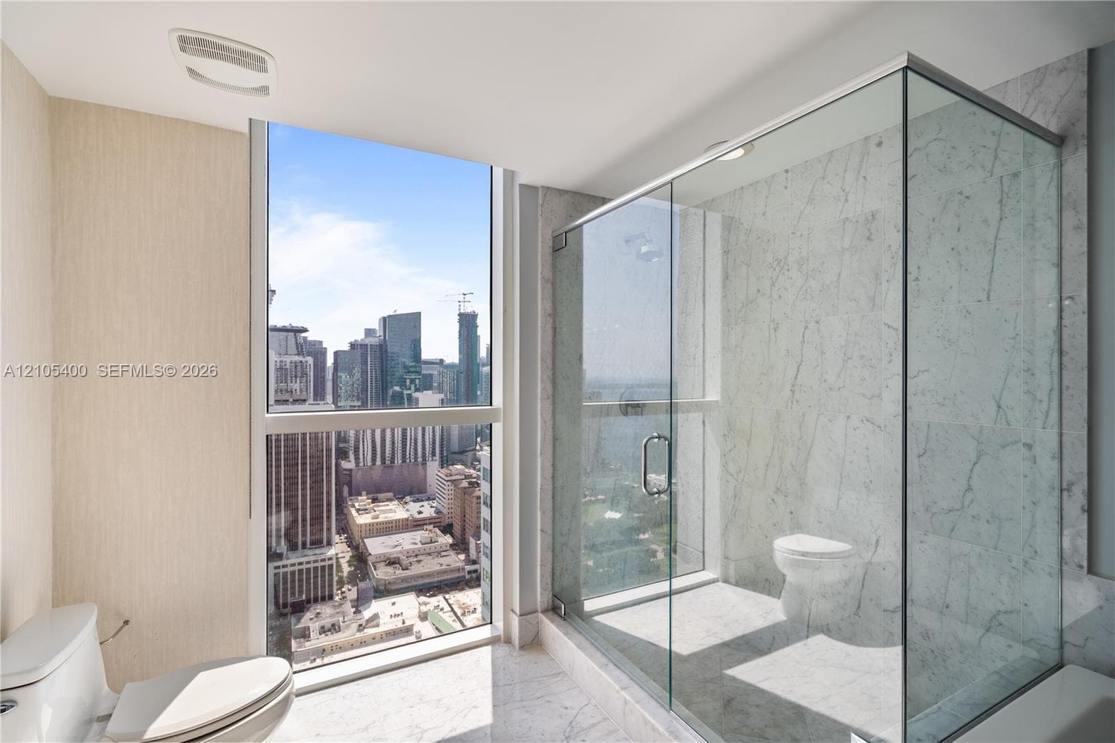 Expansive window with cityscape scenery, full glass shower enclosure, and floor-to-ceiling marbled wall and floor finishes