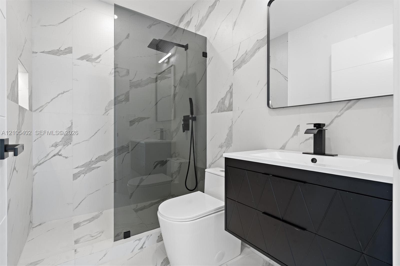 Modern bathroom featuring large-format wall and floor tile with a veined pattern, walk-in shower with glass panel, matte black plumbing fixtures, and a textured floating vanity with integrated sink