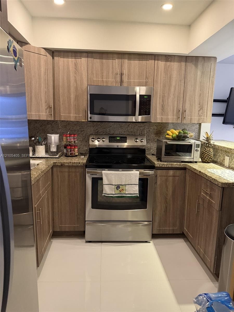 Stainless steel appliances, wood-finish cabinetry, recessed lighting, stone countertops, and a full-height stone-finish backsplash