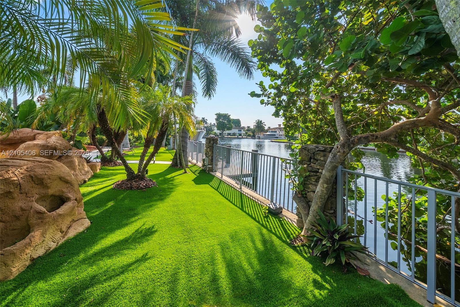 Waterfront property featuring a metal fence, lush artificial turf, mature palm trees, and decorative rock formations