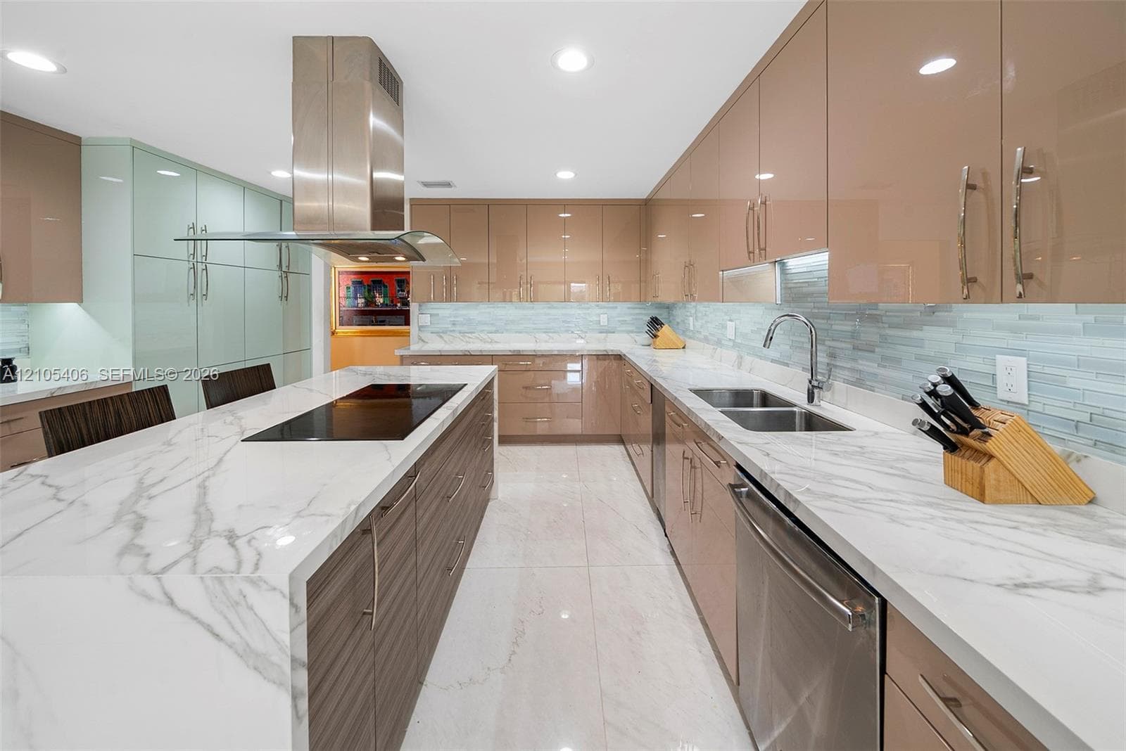 Kitchen featuring a large island with an integrated cooktop, white marble-style countertops, glossy tan cabinetry, a stainless steel range hood, and a dual basin sink