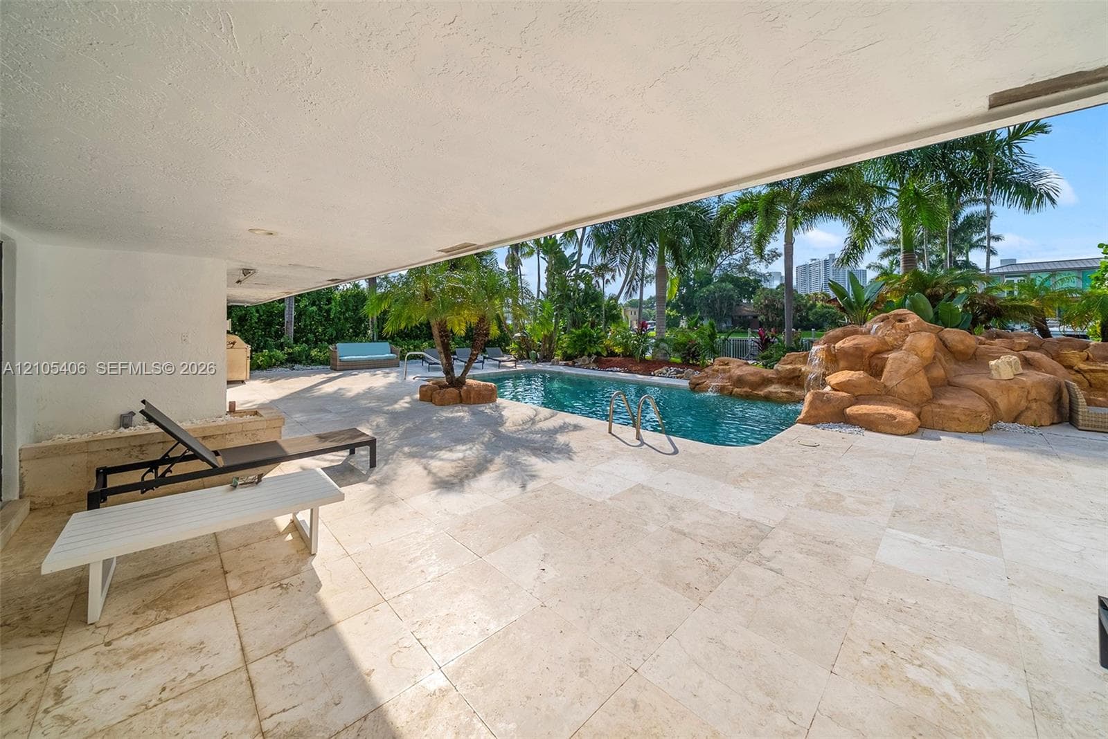 Expansive covered patio featuring light-toned tile flooring, directly accessing a custom pool with a rock waterfall feature and integrated steps