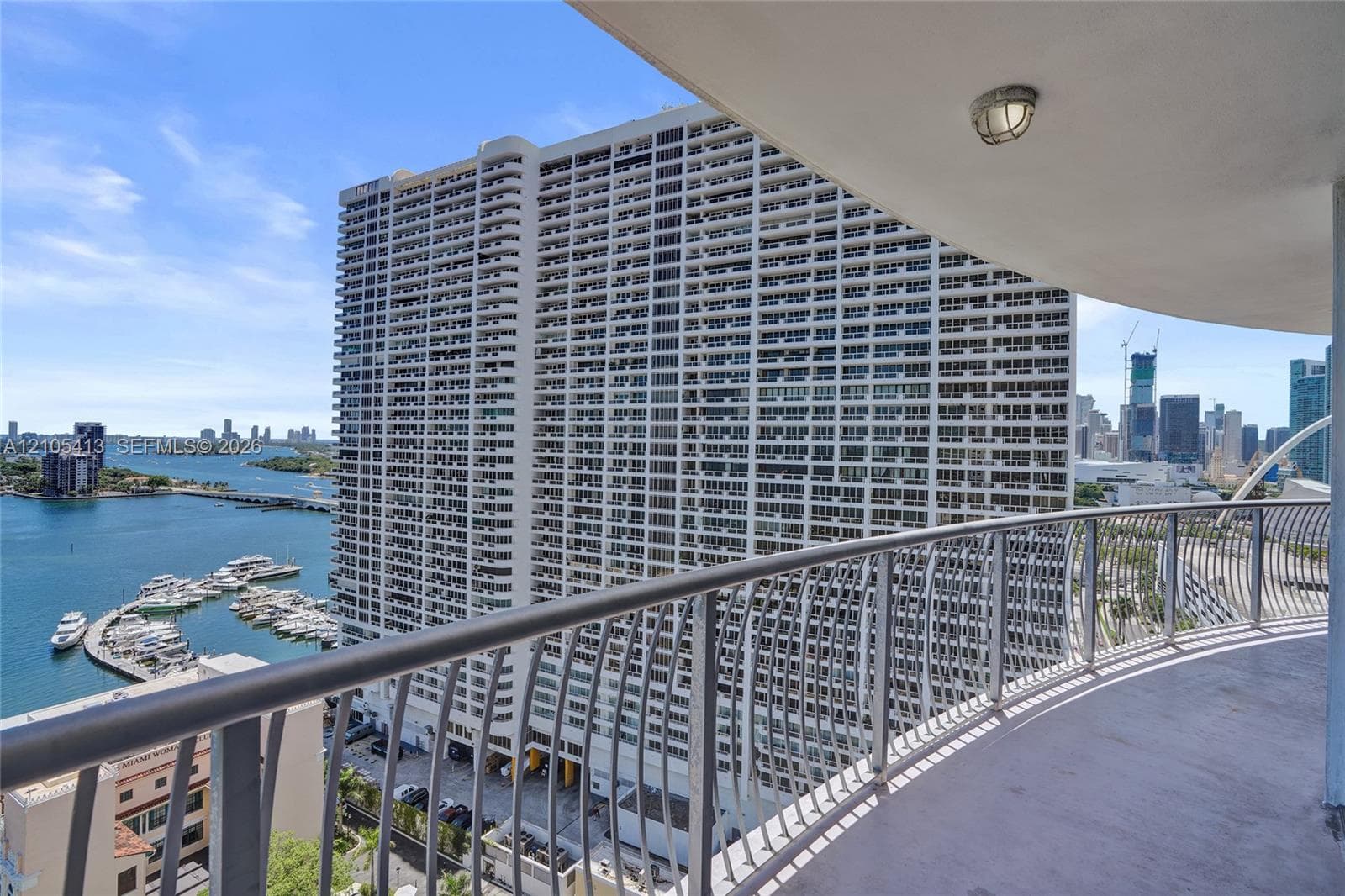 Curved balcony with metal railing, waterfront scenery, marina with multiple boats, distant cityscape, and prominent high-rise buildings