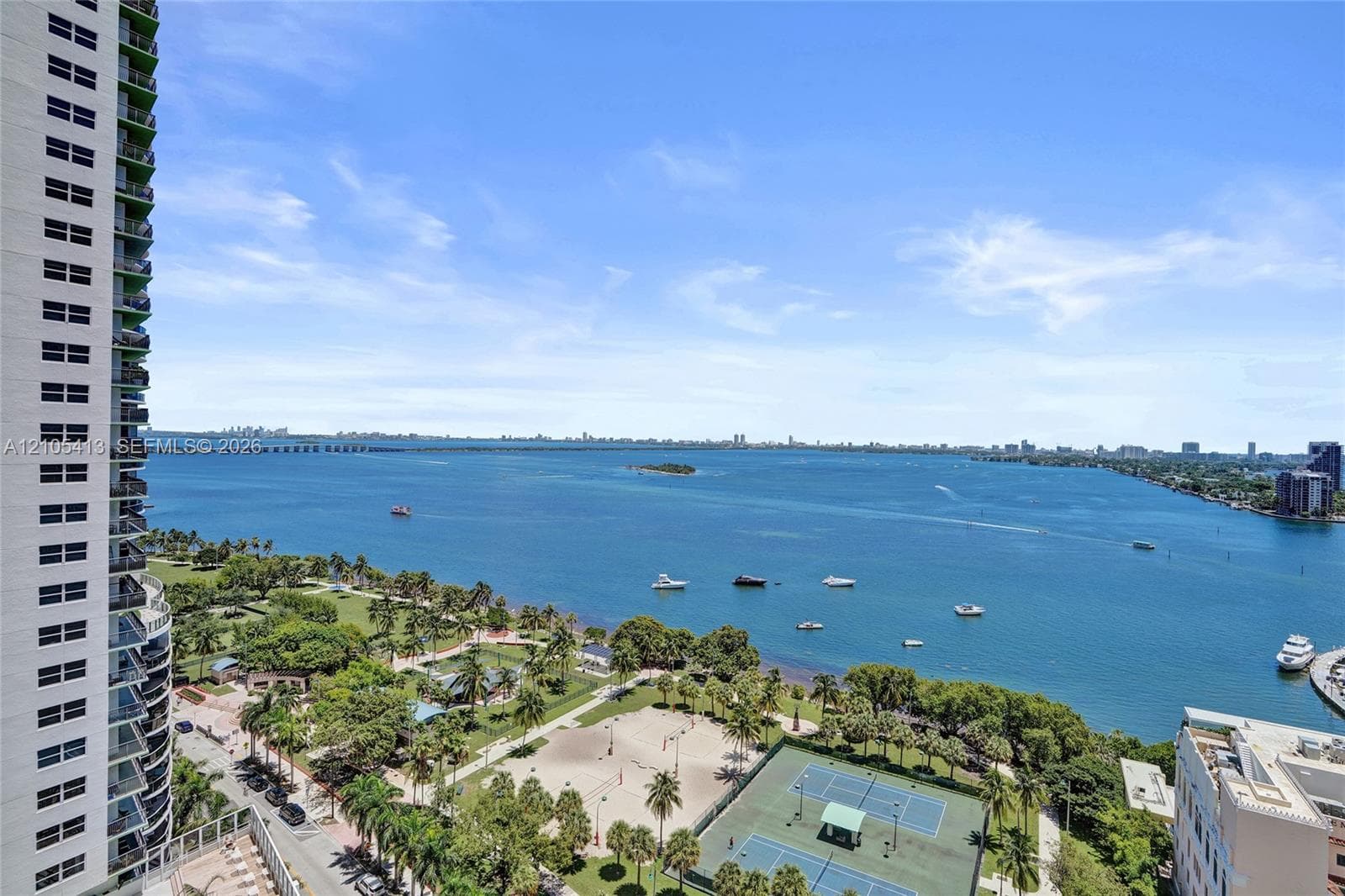 Expansive water frontage, numerous boats on the water, lush green parkland, visible tennis courts, and a distant cityscape