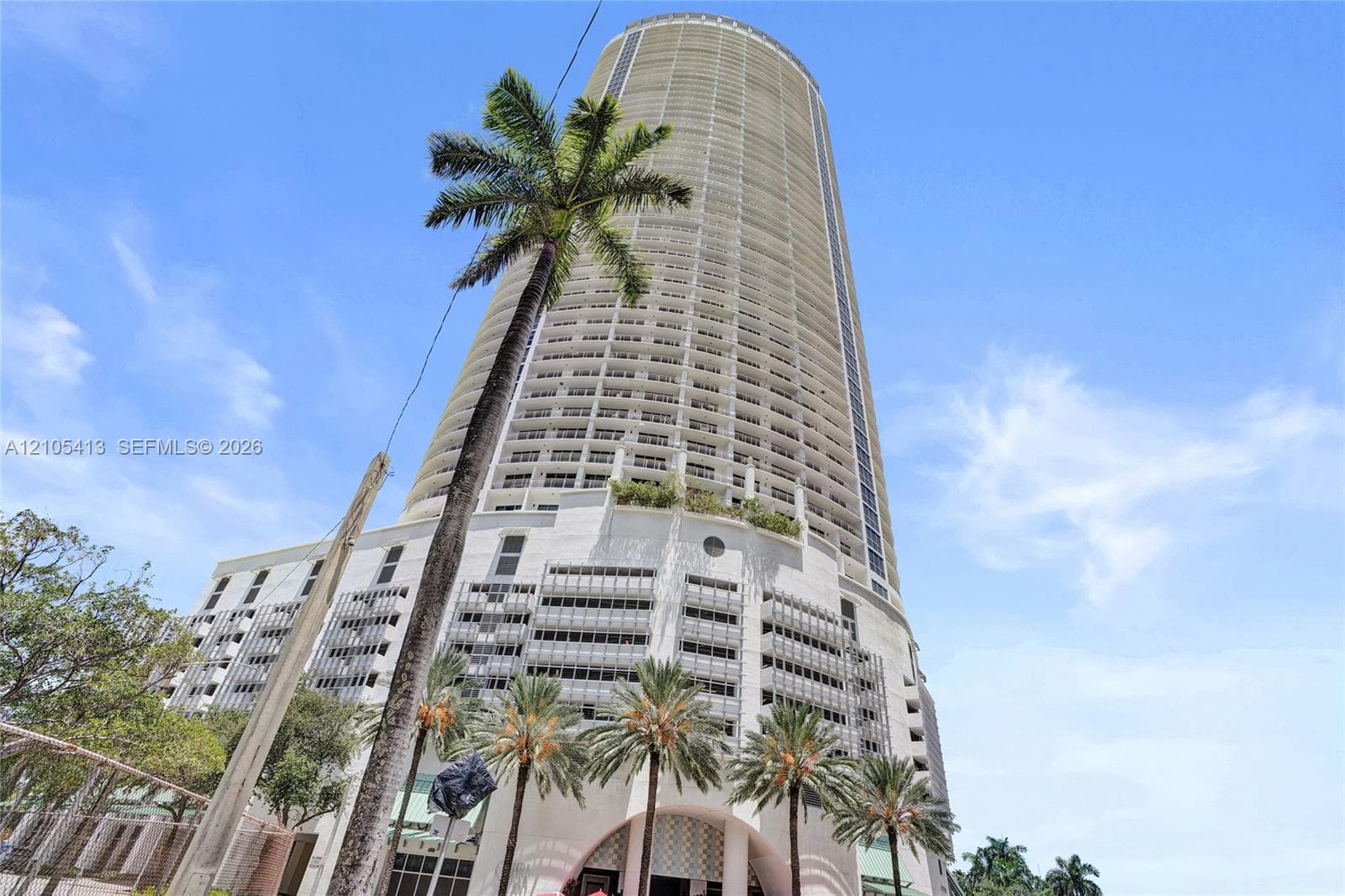 High-rise building with numerous balconies, featuring an arched entry, ground-level palm trees, and a cylindrical architectural design
