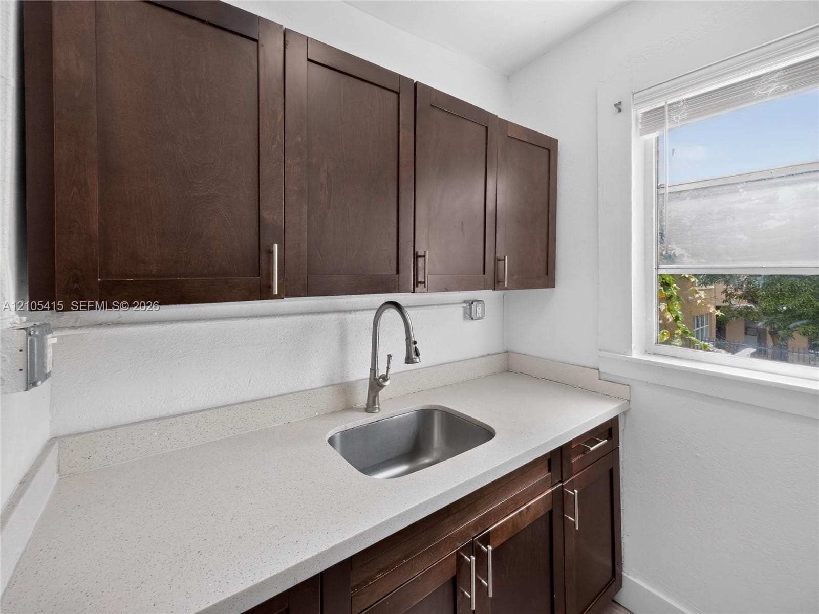 Dark wood-finish cabinetry, solid surface countertop, stainless steel undermount sink, gooseneck faucet, and window with built-in blinds