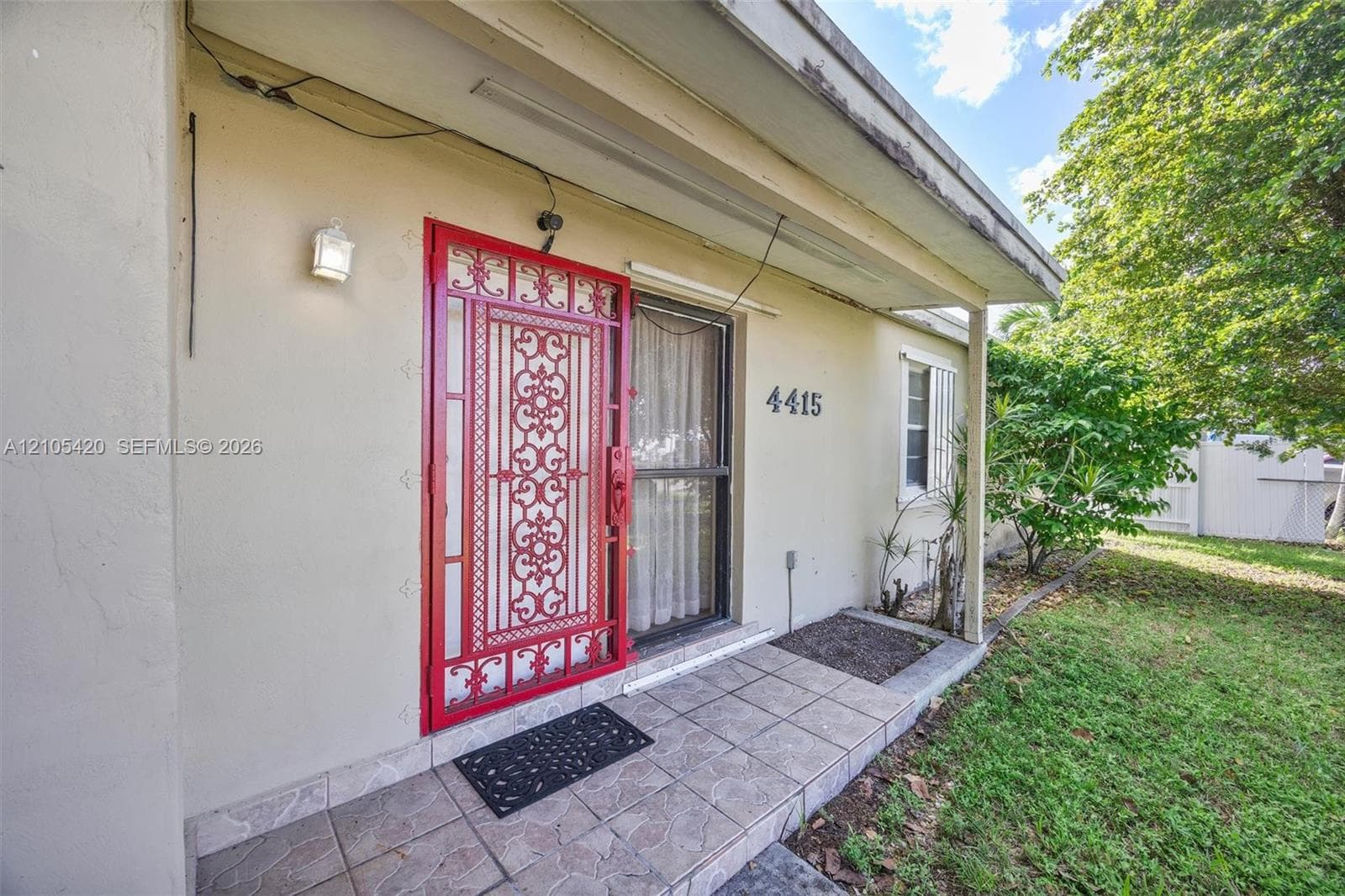 Stucco exterior featuring a red security door, wall-mounted exterior light fixture, and tiled entryway