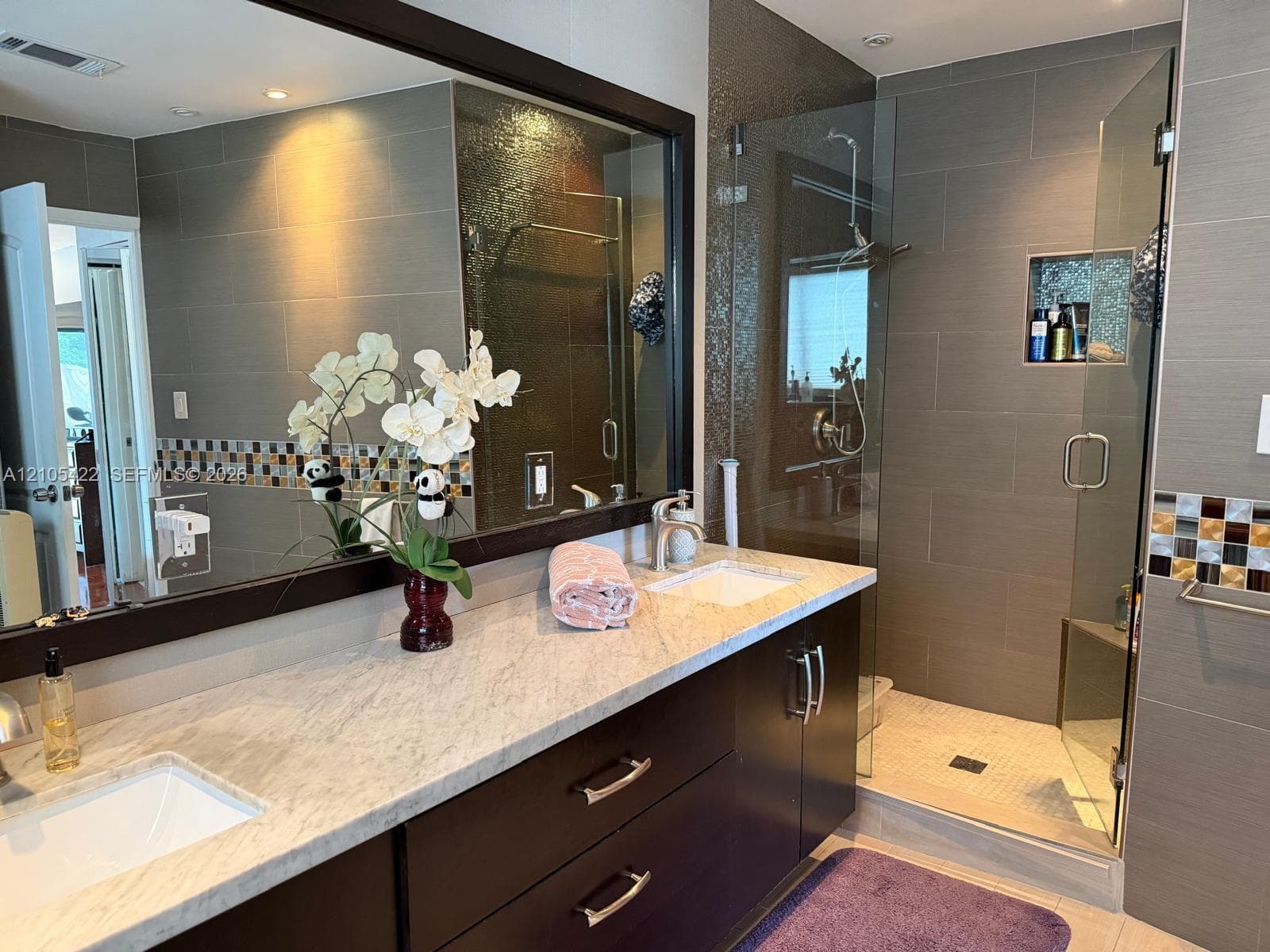 Bathroom featuring a dark wood double vanity with a stone countertop, dual undermount sinks, large framed mirror, and a walk-in glass-enclosed shower with tiled walls and a built-in shower niche
