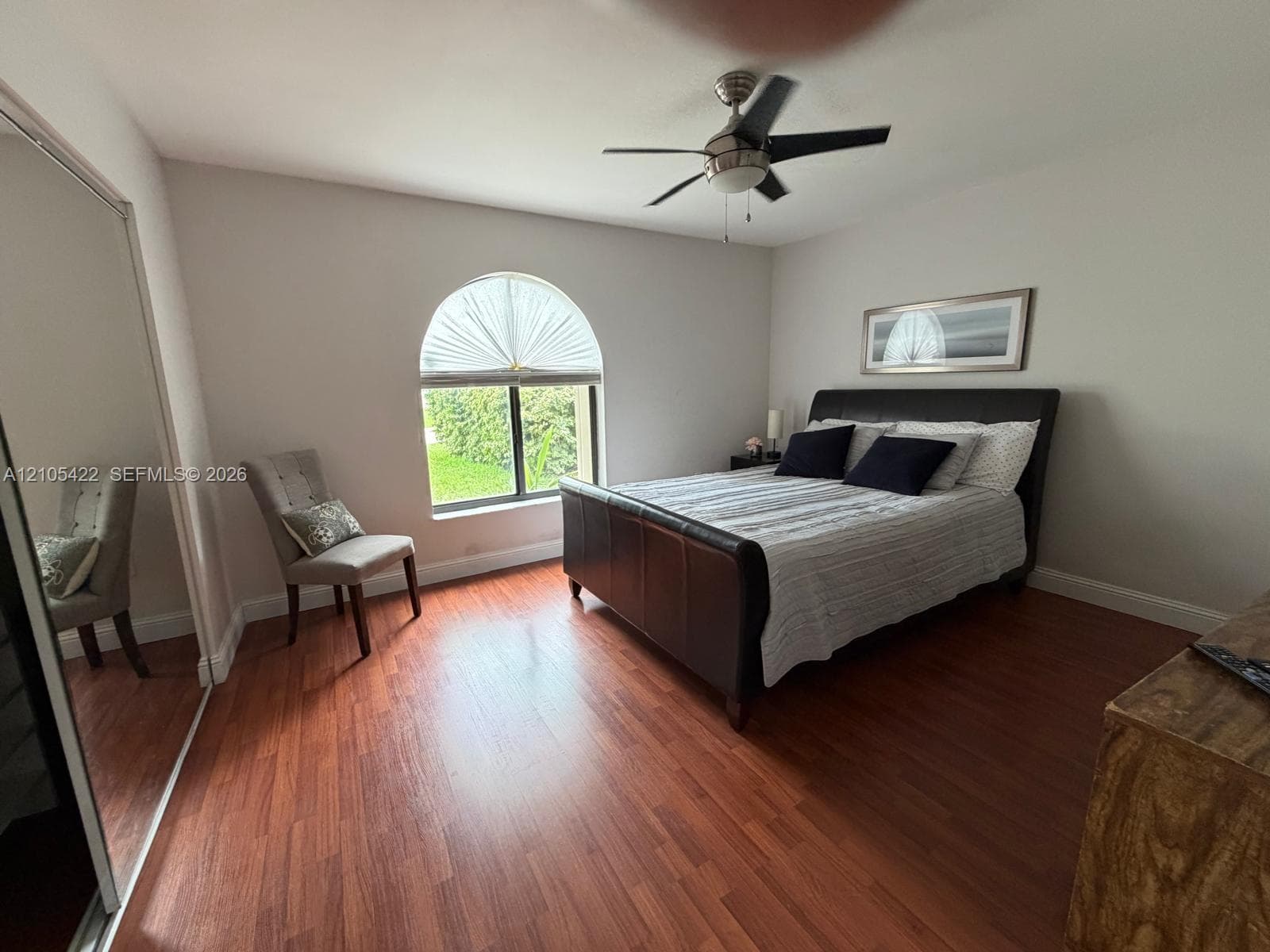 Arched window with decorative fanlight, wood-finish flooring, full-height mirrored sliding closet doors, and ceiling fan with integrated light fixture