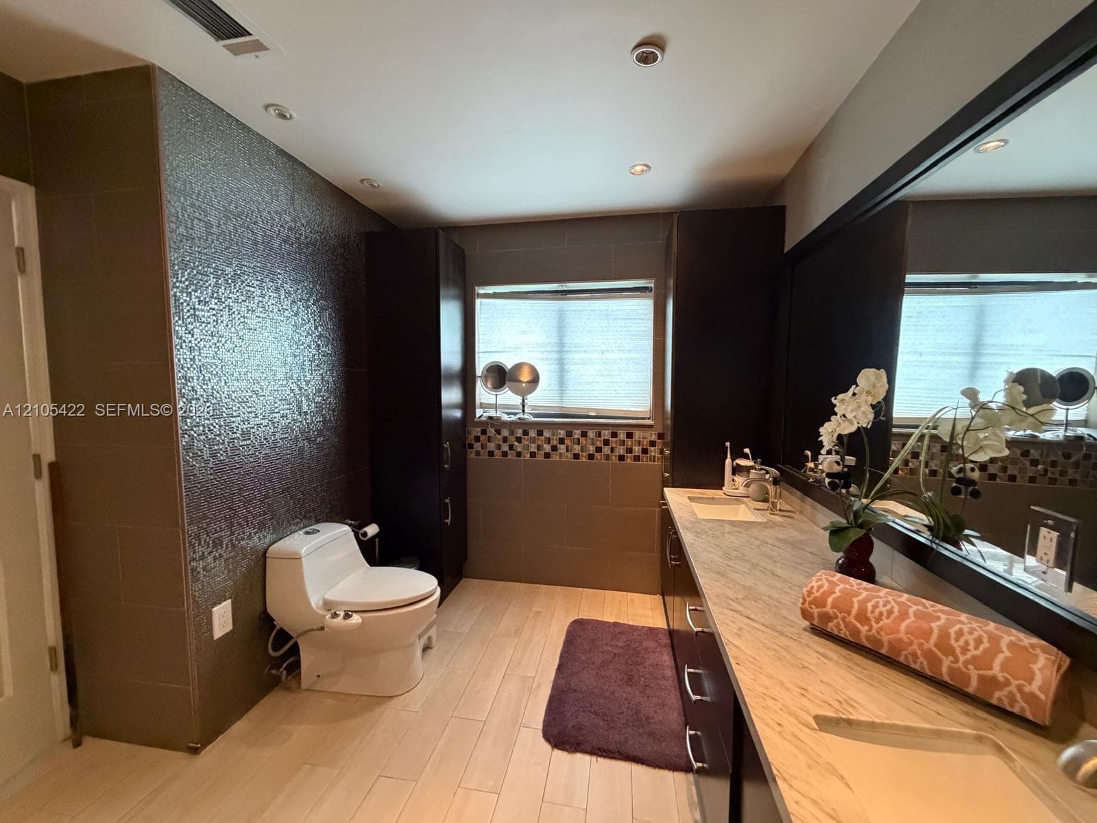 Bathroom featuring a stone-top vanity with dual integrated sinks, dark wood-finish cabinetry, floor-to-ceiling textured tile wall, and recessed ceiling lighting