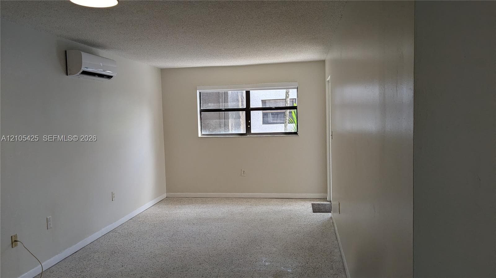 Wall-mounted air conditioning unit, light-colored walls, window with dark framing, textured ceiling, and light-colored carpeting