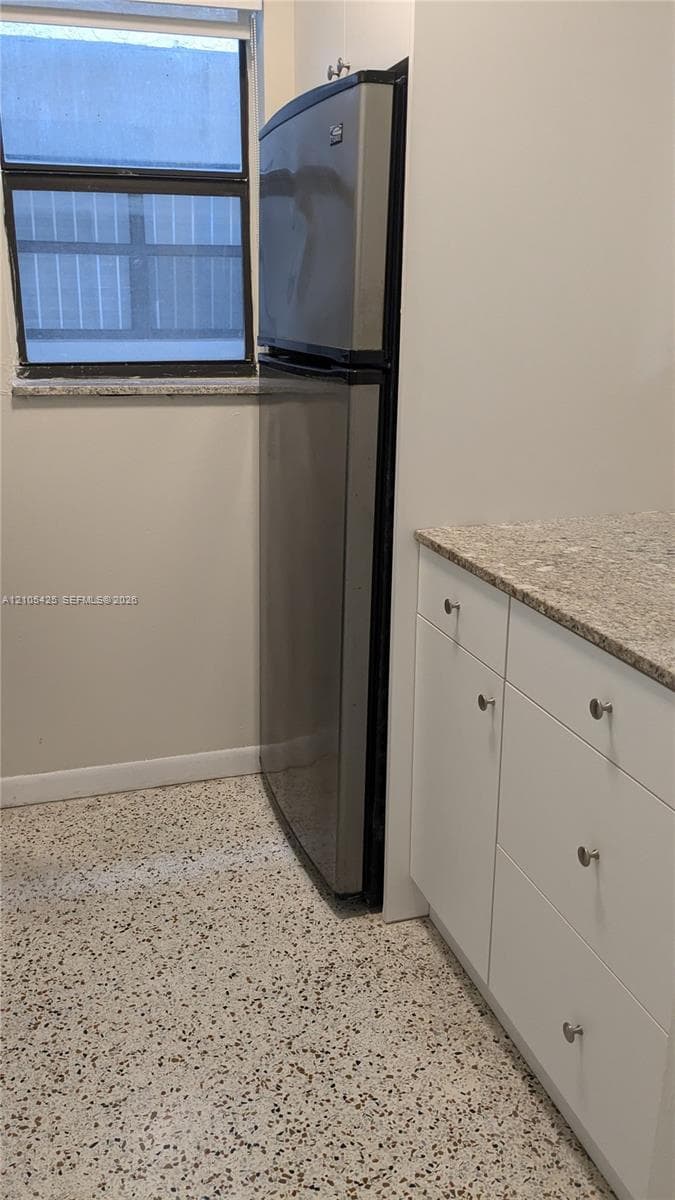 Modern kitchen featuring stainless steel refrigerator with top and bottom freezer configuration, natural stone countertops, white cabinetry with brushed metal hardware, and polished concrete flooring