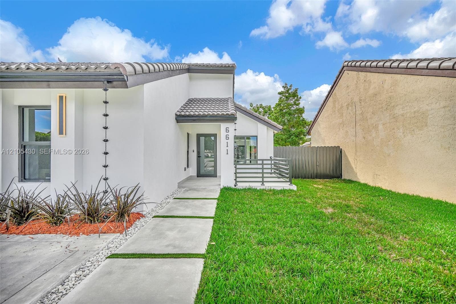 Exterior featuring smooth white stucco finish, concrete paver walkway with turf inlay, tile roof, vertical metal rain chain, and front patio with horizontal railing