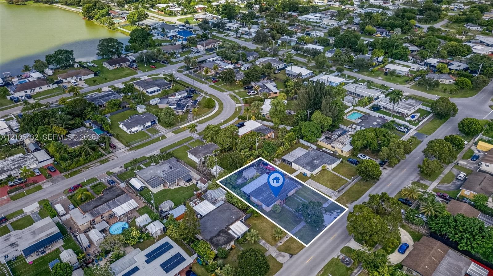 Aerial perspective of a residential lot featuring a single-story structure, expansive lawn, concrete driveway, mature landscaping, and proximity to a large body of water