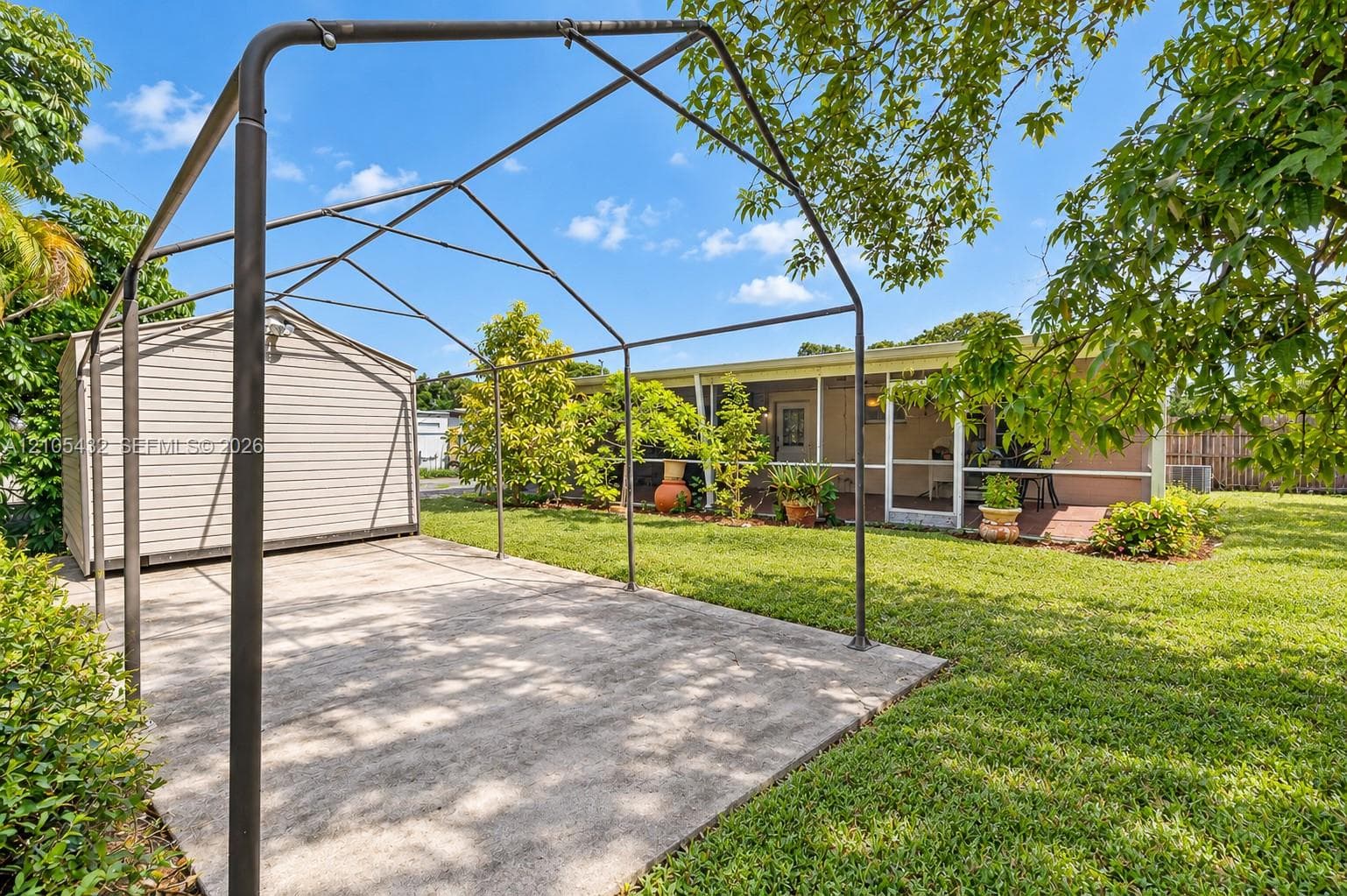 Concrete parking pad with metal frame canopy, vinyl-sided storage structure, and screened-in porch
