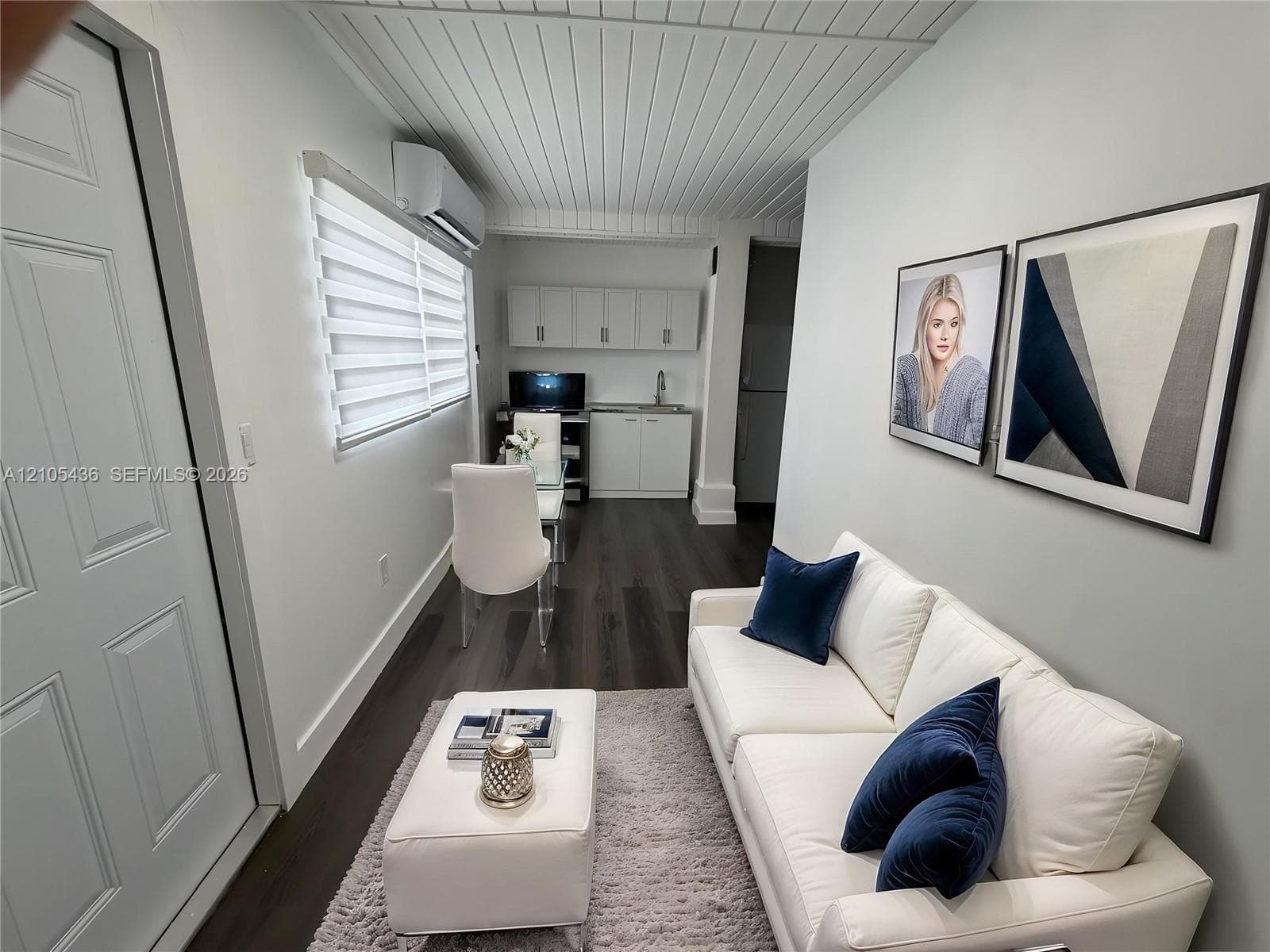 Plank-style flooring, white tongue-and-groove paneled ceiling, wall-mounted ductless heating and cooling unit, built-in white cabinetry with a sink, and window roller shades