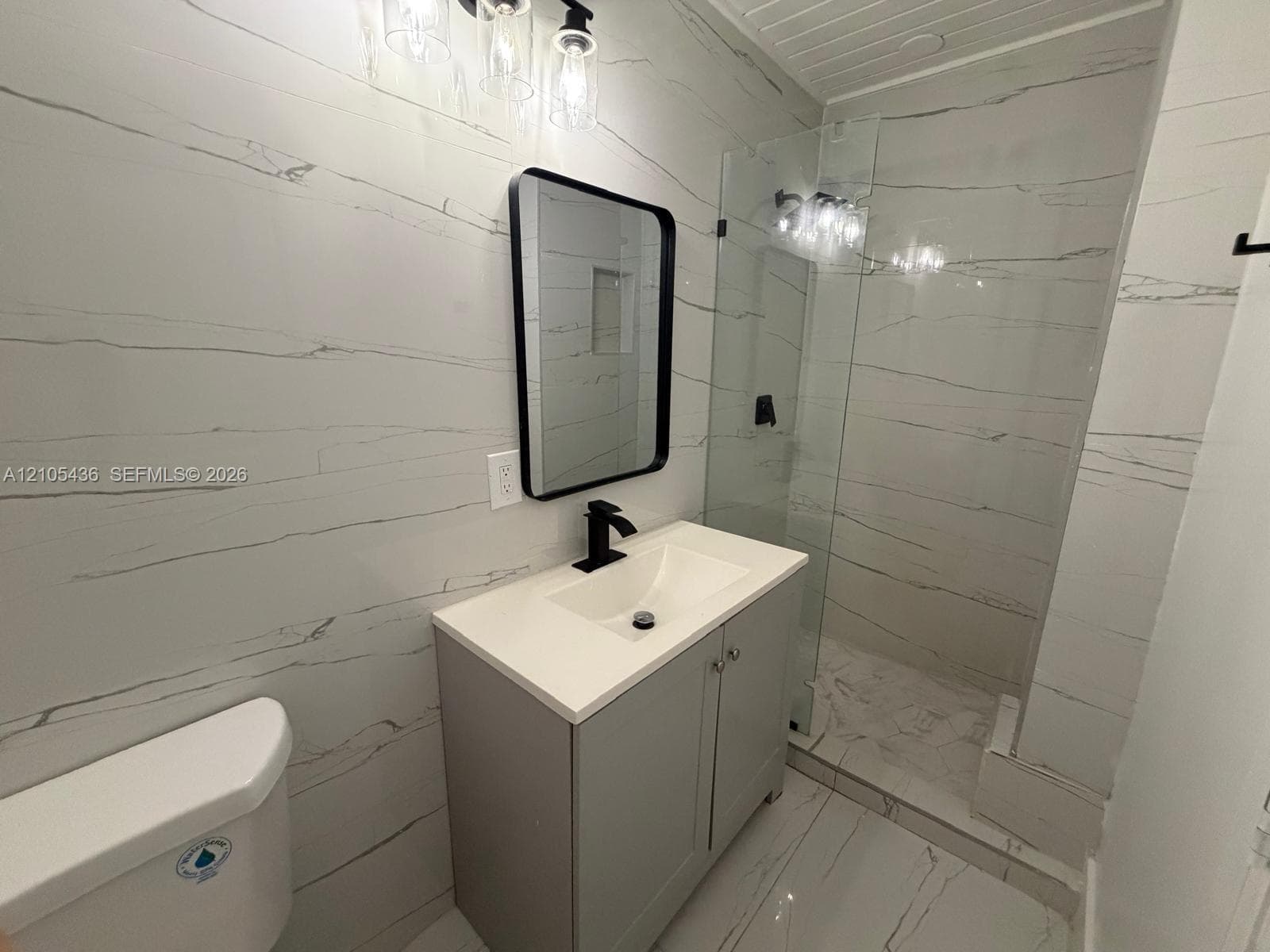 Bathroom featuring full-height stone-finish wall tile, a vanity with a solid-surface countertop, a glass-enclosed walk-in shower, matte black plumbing fixtures, and a framed mirror