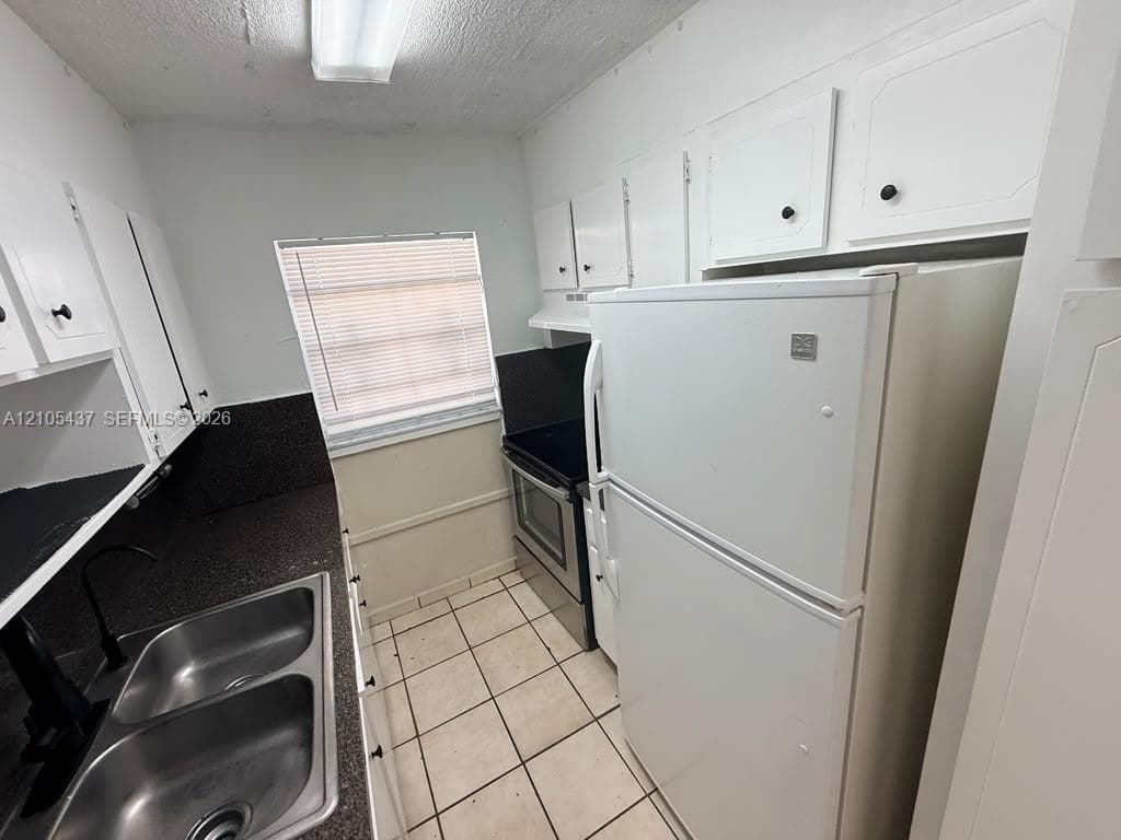 Dual basin stainless steel sink, black countertop, white cabinetry with black hardware, tile flooring, and integrated white appliances