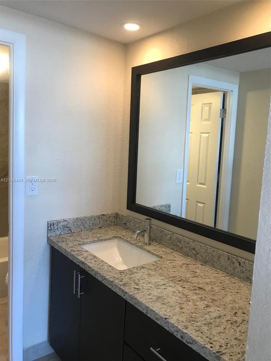 Recessed lighting, rectangular mirror with a dark frame, single basin sink, counter-mounted faucet, and dark wood-finish vanity with drawer pulls