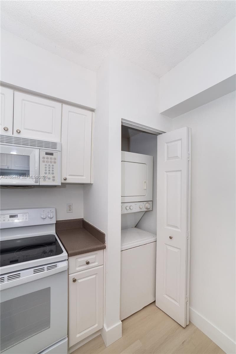 White cabinetry, white electric range, white microwave, compact stacked laundry appliances, and wood-finish flooring