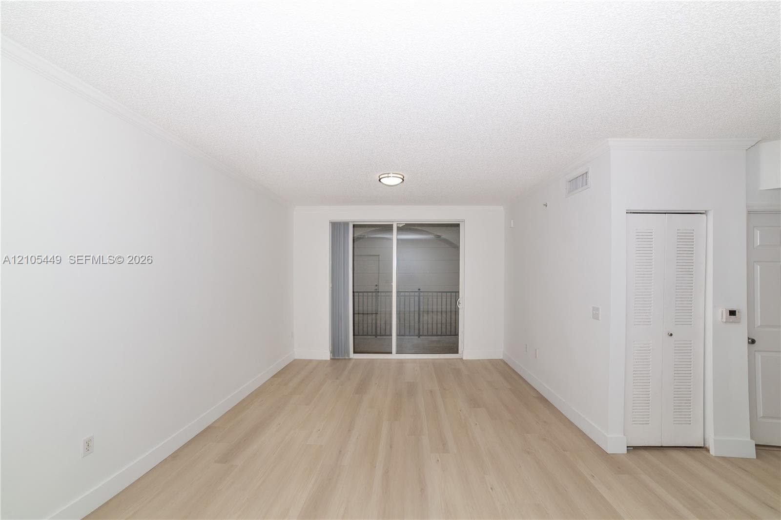 Light wood-finish flooring, sliding glass door with balcony access, crown molding, surface-mounted ceiling light fixture, and louvered bi-fold closet doors