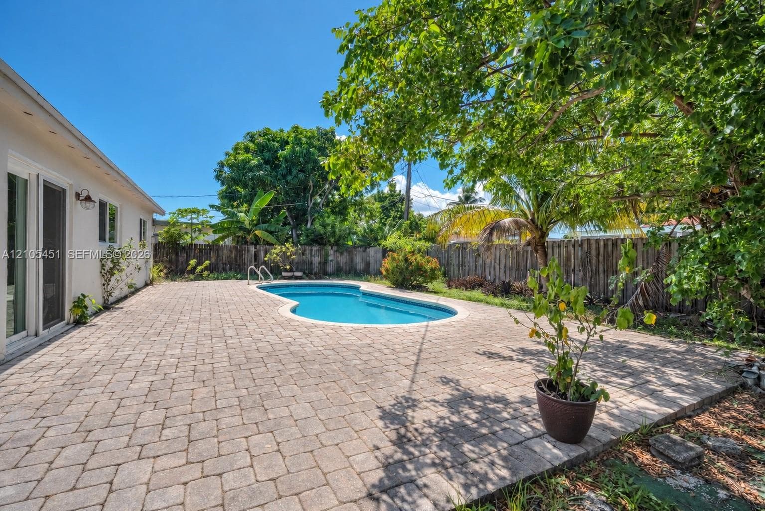 In-ground swimming pool featuring a curved design, bordered by an expansive stone-paver patio, enclosed by a wood-plank privacy fence and mature foliage